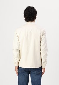 Lichtbeige jas met een kraag, lange mouwen en elastische manchetten, gemaakt van een gladde stof, gedragen over blauwe spijkerbroeken.