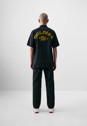 Camisa negra de manga corta con el texto y logo amarillo "GOLDIES" en la parte posterior, combinada con pantalones negros y zapatillas blancas, vista desde atrás.