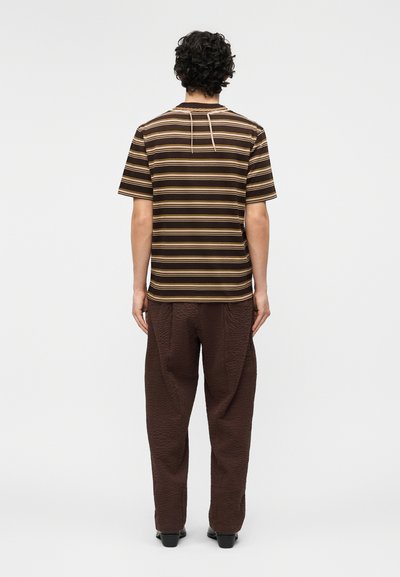 Homme aux cheveux bouclés vu de dos, portant une chemise à manches courtes marron rayée et un pantalon marron foncé texturé avec des chaussures noires.