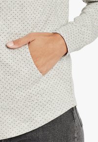 Sweatshirt gris clair avec un petit motif de points marrons, doté d'une poche latérale et de poignets côtelés. Texture douce, design décontracté et silhouette ajustée.