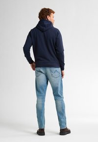 Hanorac bleumarin din material texturat, cu manșete și tiv elastic. Blugi deschiși la culoare cu un aspect uzat și cusături subtile pe buzunarul din spate. Botine maro.