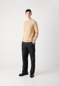 Beige kötött pulóver hosszú ujjakkal, fekete redőzött nadrággal párosítva. Fekete cipő és sapka egészíti ki az öltözetet. Egyszerű dizájn, laza fazon.