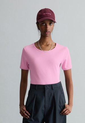 Kobieta ubrana w różową koszulkę z krótkim rękawem, ciemne plisowane spodnie, srebrny łańcuszek, kolczyki-koła oraz bordową czapkę z białym napisem.