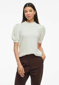 Vit texturerad blus med puffärmar och hög halsringning, tillsammans med bruna byxor. Minimalistisk design med subtilt mönster.