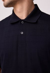 Mornarsko modra polo majica s teksturiranim karo vzorcem, z gumbno zapiralno letvijo in klasičnim ovratnikom. Gladek, zračen material.