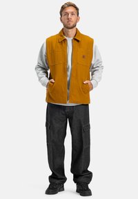 Gilet in misto cotone color senape con due tasche sul petto, abbinato a una felpa grigia e pantaloni cargo neri larghi. Design testurizzato e stratificato.