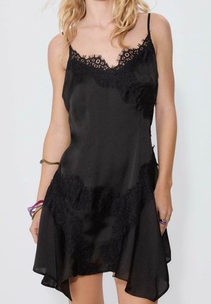 Femme portant une robe nuisette en satin noir avec bordure en dentelle, bretelles fines et ourlet asymétrique, accessoirisée de bracelets et de bagues.