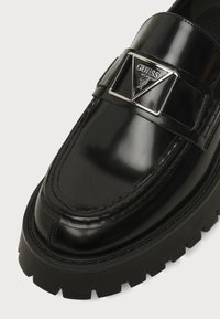 Mocassin noir brillant avec semelle épaisse à relief et une boucle triangulaire argentée avec le logo Guess sur la bride avant.