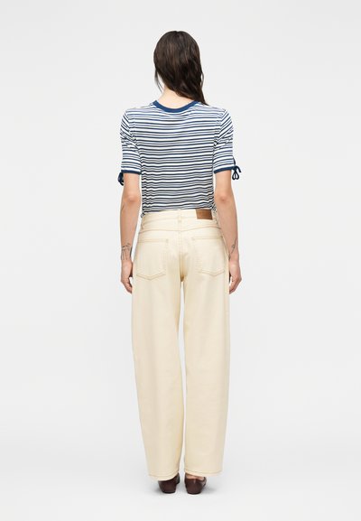 Άτομο με μακριά σκούρα μαλλιά, ντυμένο με λευκό κοντομάνικο τοπ με ναυτικό ρίγες και εκρού παντελόνι ψηλοκάβαλο με φαρδιά μπατζάκια, στέκεται μπροστά σε λευκό φόντο.