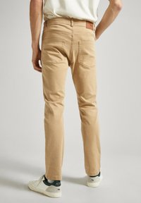 Pantalones color camel con un corte ajustado, que cuentan con bolsillos traseros y un parche de cuero marrón en la cinturilla. Combinados con zapatillas blancas.