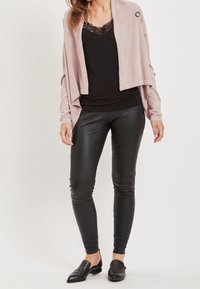 Femme portant un caraco noir à dentelle, un cardigan ouvert rose pâle, un legging en cuir noir et des mocassins noirs pointus, debout sur un fond blanc.