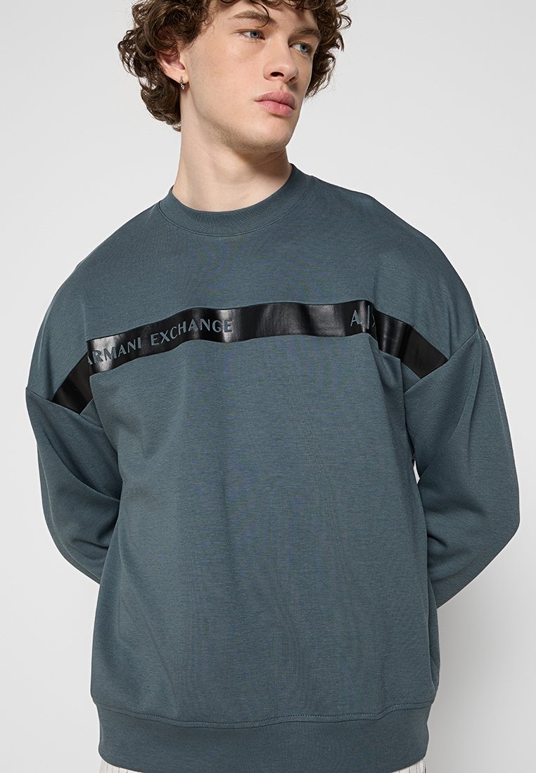Junger Mann mit lockigem Haar, der einen dunkelgrauen Armani Exchange Sweatshirt mit einem schwarzen glänzenden horizontalen Streifen über der Brust trägt.