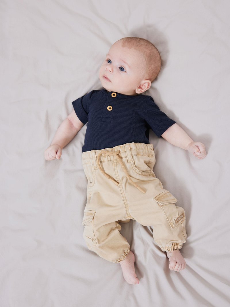 Marineblauw geribd T-shirt met korte mouwen en twee knopen aan de voorkant; beige cargo pants met elastische boorden en zakken; liggend op een lichtgekleurde vlakke ondergrond.