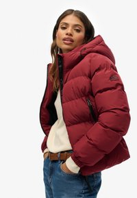 Giubbotto imbottito bordeaux con cappuccio, cerniera nera, polsini elastici e patch con logo. Indossato sopra un maglione leggero e jeans blu. Tessuto liscio e imbottito.