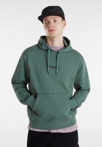 Felpa verde con cappuccio e tasca frontale, con un logo ricamato. Indossata sopra una maglietta lavanda, abbinata a un berretto nero.