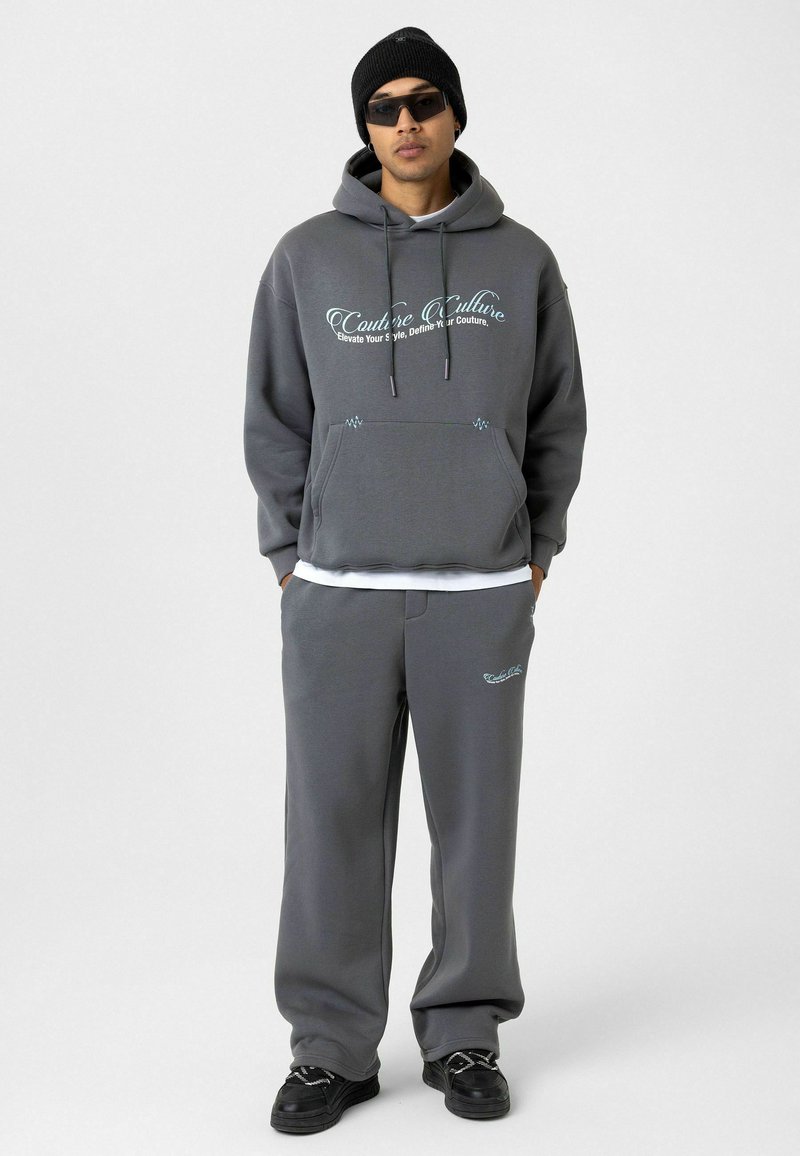 Grijze hoodie met een voorzak en trekkoorden, met bedrukte tekst. Gekoeld met grijze joggingsbroeken en zwarte sneakers. Zwarte beanie en zonnebril.