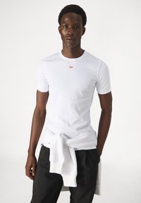 Biały, krótki rękaw, prążkowany t-shirt z okrągłym dekoltem, z małym czerwonym logo na piersi. W zestawie z luźnymi czarnymi spodniami.