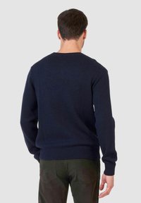 Un homme de dos portant un pull en tricot bleu marine et un pantalon vert foncé, devant un fond clair et uni.