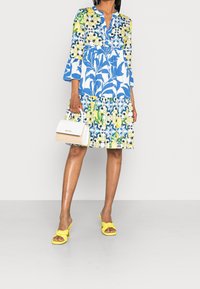 Vestido floral con patrones en azul, amarillo y verde, mangas tres cuartos y dobladillo en capas. Combinado con un pequeño bolso blanco y tacones amarillos.