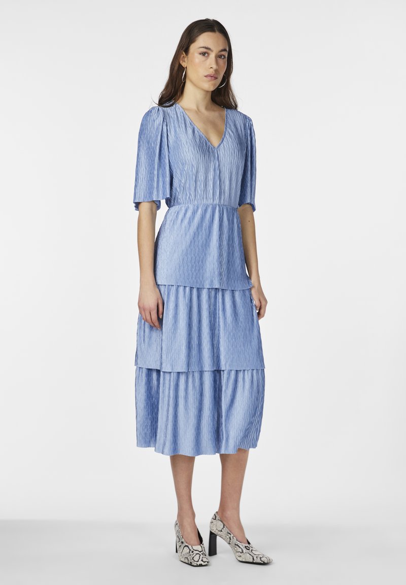 Niebieska, warstwowa sukienka z plisowanym teksturem, krótkimi rękawami i dekoltem w serek. Noszona z wzorzystymi obcasami z motywem węża.