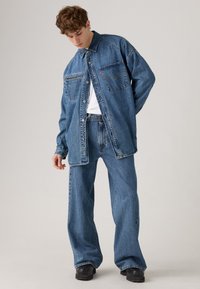 Denim jacket and wide-leg jeans in medium wash blue. Jacket features two pockets, button closure, and a collar. Black footwear.