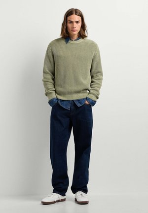 Junger Mann mit schulterlangem Haar, der einen grünen gestrickten Pullover über einem Jeanshemd, dunkle weite Jeans und weiße Sneakers trägt und vor einer hellen Wand steht.
