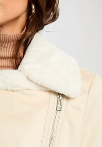 Veste en suède synthétique beige avec un col en fourrure blanche et une fermeture éclair argentée. La vue rapprochée montre la forme du col et un col roulé marron à côtes en dessous.