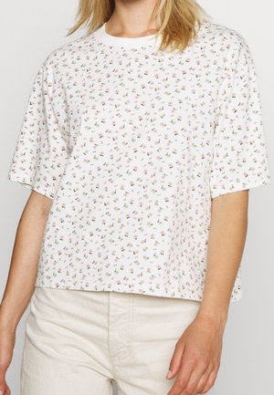 Frau trägt ein weißes, locker sitzendes T-Shirt mit einem filigranen Muster aus kleinen rosa und grünen Blumen, kombiniert mit hellen Hosen.