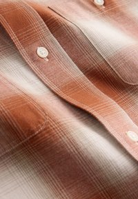 Camicia a maniche corte a quadri arancione e crema, con chiusura a bottoni, tessuto strutturato e taschino sul petto. Motivo a quadretti sottile.