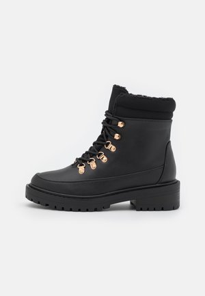 Bottine noire à la cheville avec col rembourré, crochets dorés pour les lacets, semelle épaisse à crampons et doublure en fausse fourrure, conçue pour temps froid.
