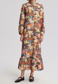 Femme portant une robe longue à manches longues, jusqu'aux chevilles, avec un motif floral coloré et des chaussures plates noires à lanières.