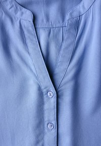 Blusa blu con scollatura a V, chiusura a bottoni e una texture di tessuto liscia. I bottoni sono rotondi con quattro fori, posti su uno sfondo chiaro.