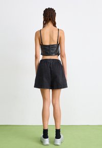 Top cropped em imitação de pele preta com alças ajustáveis, emparelhado com calções de ganga pretos de cintura alta. Modelo a usar sapatilhas prateadas e meias.