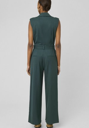 Femme portant une combinaison sans manches vert foncé à fines rayures avec jambes larges, taille ceinturée, debout devant un fond gris uni.