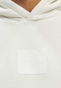 Lys grå hettegenser med en myk tekstur, som har et hvitt rektangulært merke med hevet tekst: "JACK & JONES PREMIUM EST. 1990."