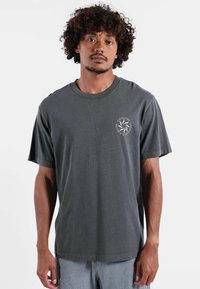 T-shirt en coton gris à manches courtes, col rond, avec un logo circulaire sur la poitrine gauche présentant un design stylisé aux accents roses.