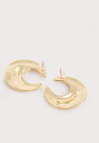 Guld halvmåneformede hoop øreringe med klare plast bagsider på en neutral overflade.