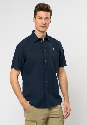 Camisa de manga corta de color marino, hecha de una tela texturizada, que presenta un cuello abotonado y un pequeño emblema en el pecho, combinada con pantalones cargo beige.