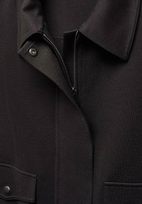 Gros plan sur l'avant d'une veste noire montrant le col, la fermeture éclair partiellement ouverte, le bouton-pression et la poche à rabat avec fermeture par bouton.