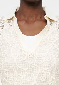 Κοντινό πλάνο ενός ατόμου που φοράει κρεμ τοπ με δαντέλα στο γιακά πάνω από λευκή φανέλα, εμφανίζοντας περίπλοκα ανθικά σχέδια στο ύφασμα.