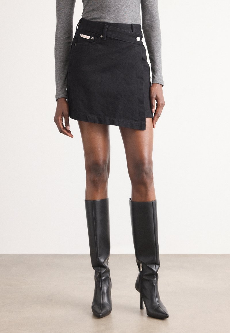Femme portant un haut gris à manches longues, une mini-jupe asymétrique noire et des bottes noires hautes à talons jusqu'aux genoux, debout sur un sol plat.
