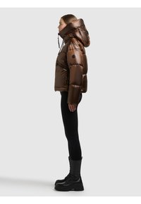 Brązowa kurtka puchowa z błyszczącym wykończeniem, oversize'owym kapturem i kieszeniami na zamek, noszona z czarnymi legginsami i wojskowymi butami, widziana z boku.