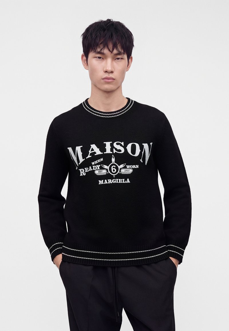Giovane uomo con capelli neri indossa un maglione nero con testo bianco "Maison Margiela" e grafica, mani nelle tasche, sfondo neutro.