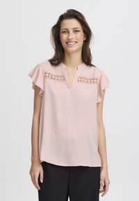 Hellrosa Bluse mit kurzen Rüschensleeves, V-Ausschnitt und dekorativen Spitzeing Details über der Brust. Glatter Stoff und lockerer Schnitt.