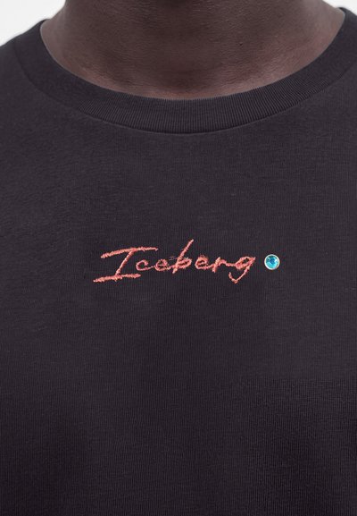 Крупен план на черна риза с червенизбродирано "Iceberg" текст и малък син декоративен камък близо до врата.