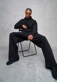Sudadera negra de gran tamaño con cuello alto y pantalones negros holgados, sentado en una silla minimalista de metal negro contra un fondo gris.
