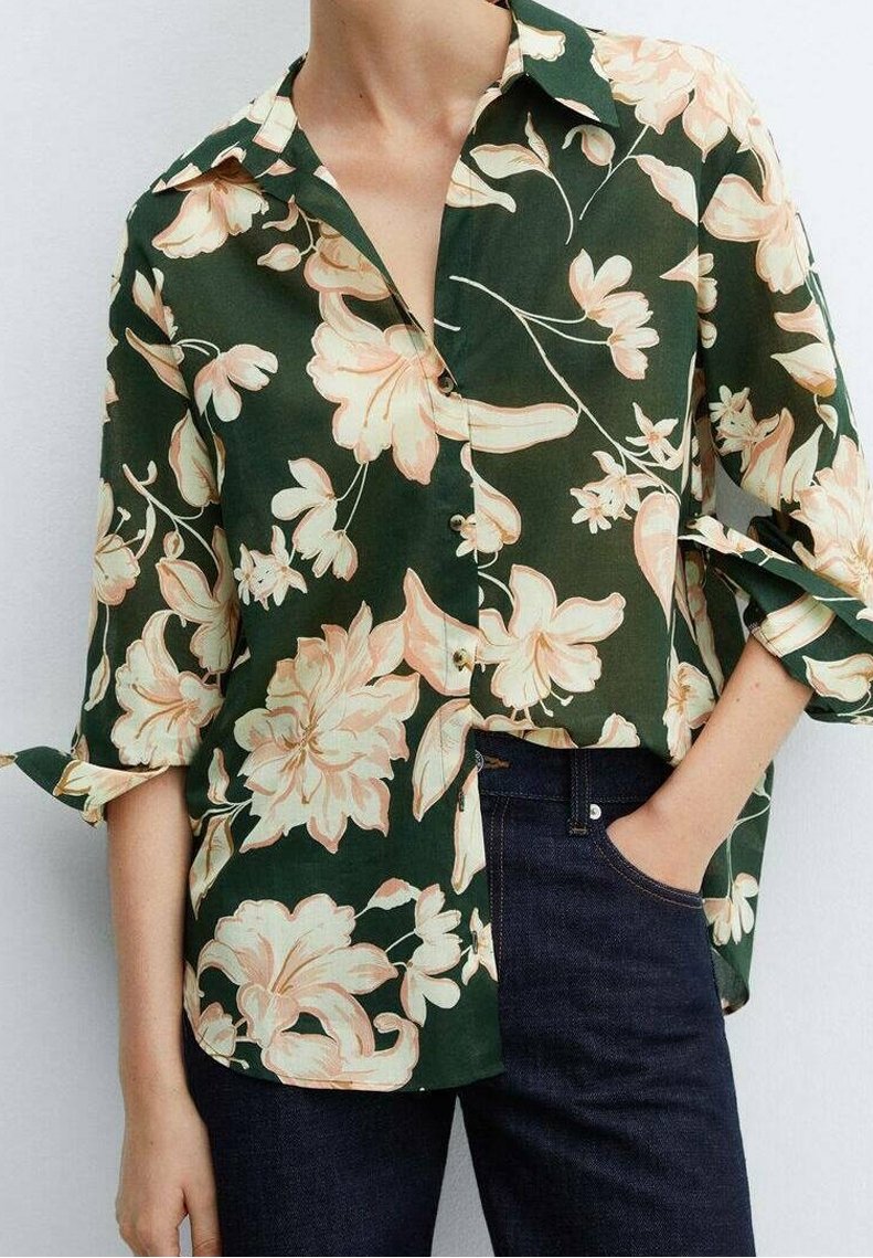 Personne portant une chemise boutonnée vert foncé avec un imprimé floral beige et un jean bleu foncé, une main dans la poche.