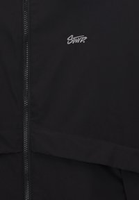 Veste noire avec fermeture éclair à l'avant et petit logo brodé blanc "SWT ?" sur la partie supérieure de la poitrine.