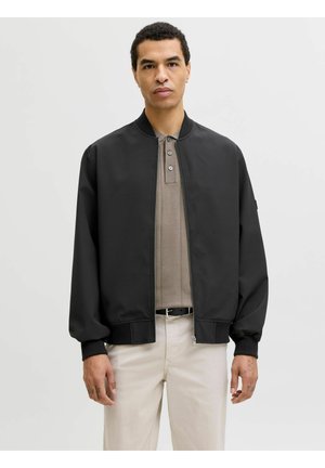 Mann trägt eine schwarze Bomberjacke über einem taupefarbenen Poloshirt und beige Hose, steht vor einem schlichten, hellen Hintergrund.