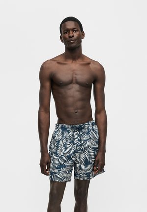 Λεπτός άνδρας με σκουρόχρωμο δέρμα όρθιος, φοράει μπλε μαγιό σορτς με λευκά μοτίβα φύλλων φοινικόδεντρου, σε απλό ανοιχτό γκρι φόντο.
